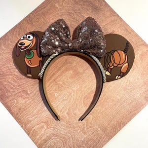 May include: Brown mouse ears headband featuring Slinky Dog and a sequined bow. The ears have a brown background with Slinky Dog illustrations. The headband is brown with a silver rope trim.
