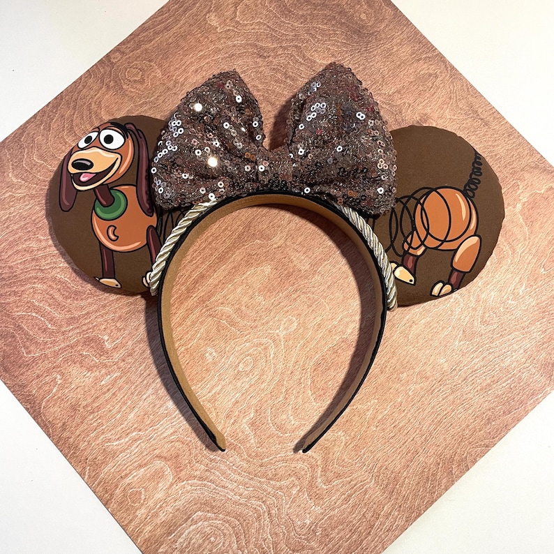 May include: Brown mouse ears headband featuring Slinky Dog and a sequined bow. The ears have a brown background with Slinky Dog illustrations. The headband is brown with a silver rope trim.