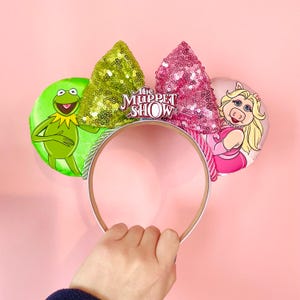 May include: A headband featuring two circular ears with illustrations of Kermit the Frog on the left and Miss Piggy on the right. Between the ears are two sequined bows in lime green and pink, with a central emblem reading The Muppet Show.