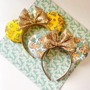May include: Three headbands with mouse ears. The ears feature a yellow background with a lion cub design and the text "Little King". The headbands have gold sequin bows and gold rope trim.