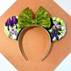 May include: A headband featuring two circular ears with a green and white diagonal striped pattern. The left ear displays a cartoon character with a purple bow, and the right ear shows a cartoon character in a dark jacket. A large green sequined bow sits between the ears.