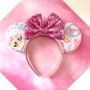 Op de afbeelding: Roze pailletten strik haarband met muizenoren. De oren zijn lichtroze met een patroon van stripfiguren en iconen. De haarband is zilverkleurig en de strik is bedekt met roze pailletten.