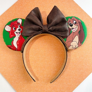 May include: A headband featuring two circular green ears with cartoon dog illustrations. The left ear shows a red dog, and the right ear shows a brown dog. A large, textured dark brown bow sits between the ears, and the headband has a twisted rope trim.
