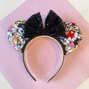 Può includere: Fascia per capelli con orecchie da topo, con un fiocco nero con paillettes e orecchie decorate con personaggi dei cartoni animati. Le orecchie mostrano Topolino, Minnie e Paperino. La fascia è nera e beige.