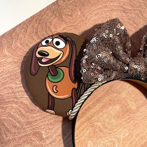 May include: Brown mouse ears featuring a cartoon image of Slinky Dog. The ears have a large, sequined bow in shades of brown and gold. The headband is trimmed with a gold and white rope.