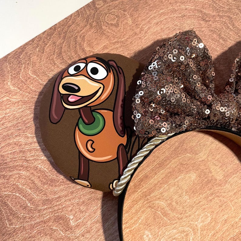 May include: Brown mouse ears featuring a cartoon image of Slinky Dog. The ears have a large, sequined bow in shades of brown and gold. The headband is trimmed with a gold and white rope.