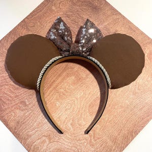 May include: Brown mouse ear headband with a sequined bow. The ears are a solid brown color, and the bow is made of shimmering sequins. The headband is black with a silver rope trim.