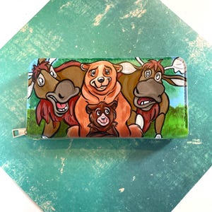 Peut inclure: Un portefeuille coloré avec une illustration de dessin animé d'une famille d'ours et de deux élans. Le portefeuille a une fermeture éclair et est peint avec des couleurs vives. Le fond est un paysage vert et bleu.