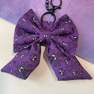 May include: A fabric bow keychain featuring a purple background with a repeating black and white pattern of stylized ghost-like figures and swirling shapes. The bow is attached to a black metal split ring and a lobster claw clasp hardware.