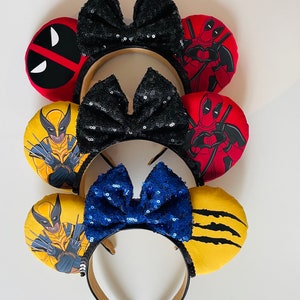 May include: Three pairs of character-themed mouse ears with black, blue, and yellow fabric. The ears feature images of Wolverine and Deadpool. The bows are black, blue, and black with sequins.