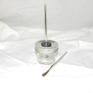 Op de afbeelding: Een glazen pot met een zilveren deksel en een zilveren metalen roerstok. De pot heeft een diamantpatroon aan de zijkanten. Een zilveren metalen borstel ligt op het witte oppervlak.