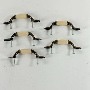 May include: Five drawer pulls featuring a central cream-colored ceramic handle section flanked by curved, dark bronze-toned metal ends. Each pull includes two silver-colored metal mounting screws extending downward from the base of the hardware.
