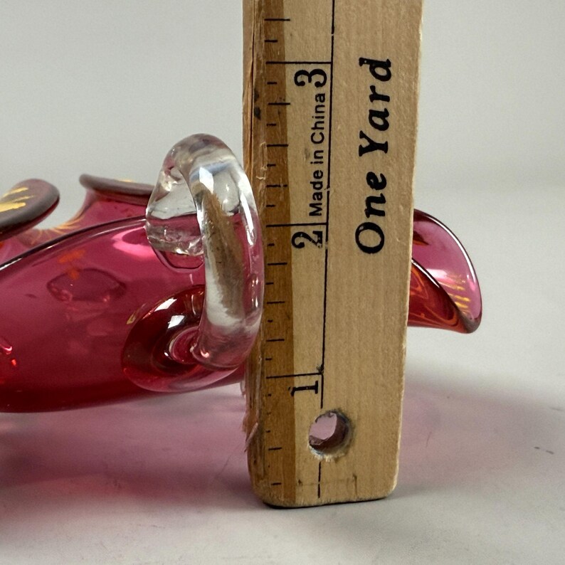 May include: A pink glass candy dish with a clear glass handle. The dish is about 2 inches tall. "Made in China" and "One Yard" are printed on a wooden ruler next to the dish.