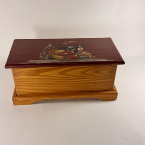 May include: A rectangular wooden box with a natural light brown wood grain finish on the sides and base. The lid is painted a deep maroon color and features a detailed illustration of a bowl of apples, a glass lantern, and a lit candle.