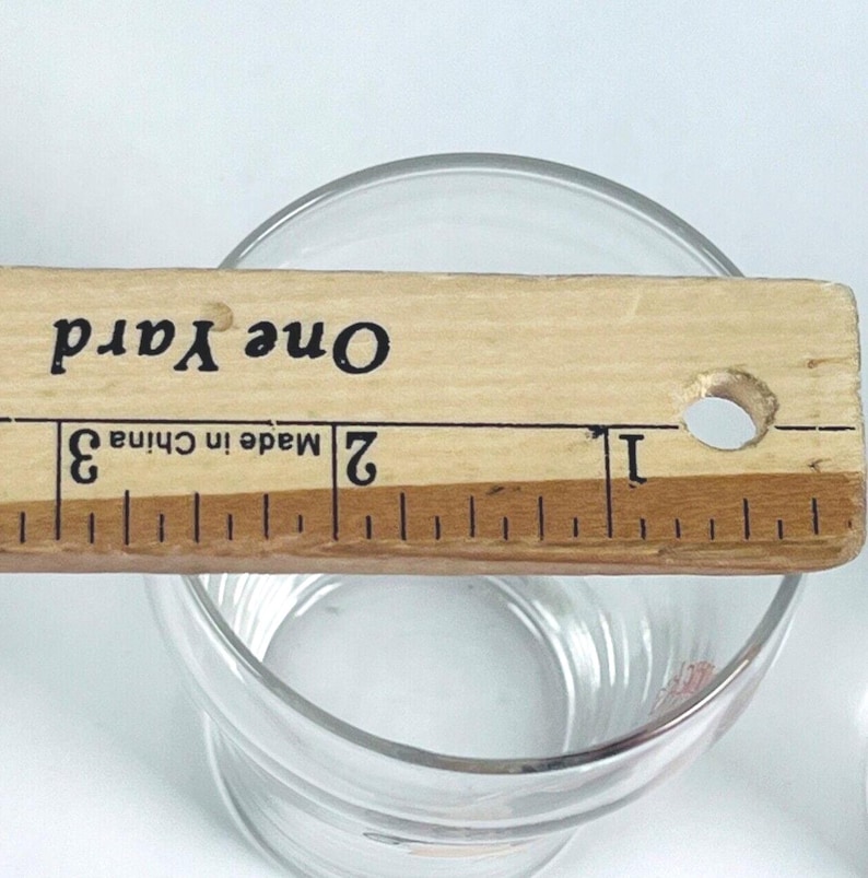 May include: A clear glass tumbler with a wooden ruler placed across the top. The ruler is marked with inches and the text "One Yard" and "Made in China". The tumbler is on a white surface.