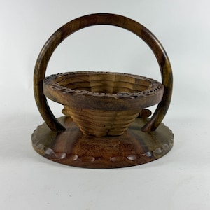 May include: A dark brown wooden basket featuring a circular base and a tall, arched handle. The basket has a carved, textured rim and a deep, bowl-shaped interior with visible wood grain patterns throughout the entire structure.