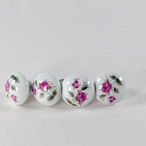 May include: Four round white ceramic cabinet knobs, each featuring a hand-painted design of pink rose blossoms with green leaves and stems. The knobs have a smooth, glossy finish and a metallic mounting hardware piece visible on the back.