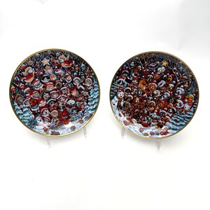 May include: Two decorative plates with a gold rim. One plate features a crowd of Santa Clauses, and the other plate features a crowd of dogs in winter attire. The plates have a winter holiday theme.