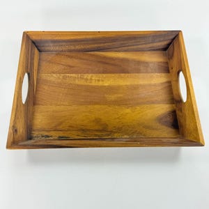May include: A rectangular wooden serving tray featuring a natural brown wood grain pattern. The tray has raised sides with integrated circular cut-out handles on the left and right ends, providing a functional grip for carrying items securely.