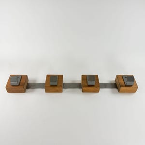 May include: A wall-mounted coat rack featuring a horizontal metal bar connecting four square wooden blocks. Each wooden block is fitted with a single, angled, brushed metal hook. The design is linear, combining natural wood tones with industrial metal elements.