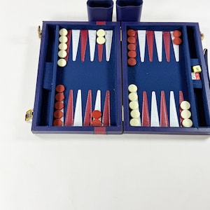 May include: A blue backgammon board set featuring a dark blue felt interior with alternating white and maroon triangular points. The set includes cream and maroon circular checkers, two blue dice cups, and dice showing the number eight and other pips.
