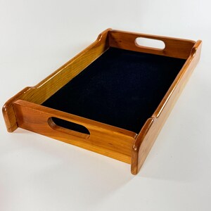 May include: A rectangular wooden serving tray featuring a polished light brown wood grain finish and a smooth black felt-lined base. The tray includes two integrated cutout handles on the shorter sides, providing a functional design for carrying.