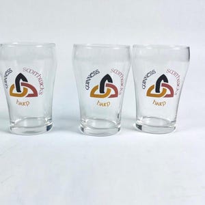 May include: Three clear pint glasses, each bearing the Guinness logo with a harp design and the words "SCOTTUCKS" and "HARP". The glasses are empty and stand on a white surface.