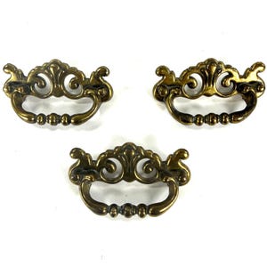 May include: Three antique brass drawer pulls with ornate, scrollwork designs. Each pull features a curved handle with a series of small, decorative beads. The pulls have a warm, aged patina, suggesting vintage or retro styling. Perfect for furniture restoration.