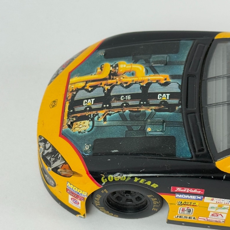 May include: A detailed close-up of a toy race car. The car is primarily yellow and black, with the words "Good Year" on the tire. The hood features a graphic of an engine with the "CAT" logo. Additional logos and text are visible on the car's body.