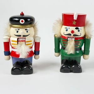 May include: Two wooden nutcrackers, one in a black hat with a red and white uniform, and the other in a red hat with a green uniform. Both have white hair and mustaches, and black boots. They are standing on a white surface.