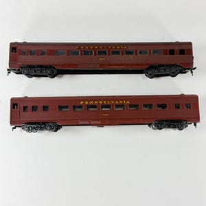May include: Two maroon model train passenger cars with black undercarriages and wheels. Each car features the word PENNSYLVANIA in yellow lettering along the top side, with one car marked 4203 and the other marked CLEAR below the name.