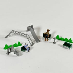 May include: Miniature playground set with a gray slide, ladder, and seesaw. Includes small green bushes, benches, and figures of people. The set is designed for a model train or diorama.