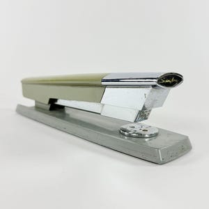 May include: A vintage Swingline stapler in shades of silver, chrome, and olive green. The stapler features a sleek, streamlined design with the Swingline logo in gold script. The base measures approximately 6 inches in length.