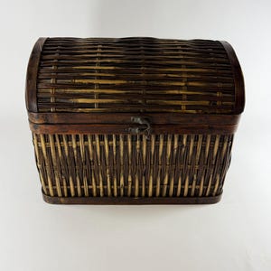 May include: A dark brown woven wicker storage chest with a rounded, arched lid. The chest features a vertical weave pattern on the body and a horizontal weave on the lid, secured by a small, centered metal latch on the front edge.