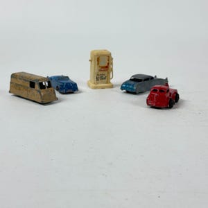 May include: A collection of vintage toy vehicles, including a red truck, a blue car, a gray car, a tan van, and a cream-colored gas pump. The gas pump has the text "Hi-Test" on it. The toys are arranged on a white surface.