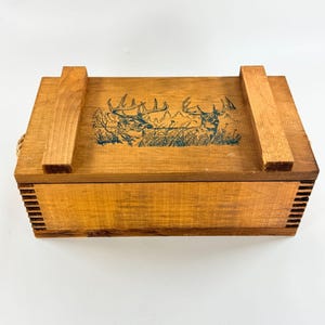 May include: A rectangular wooden box with a natural light brown finish and finger-jointed corners. The lid features a dark green graphic illustration of two deer standing in a grassy field. Two raised wooden slats are attached to the top lid.