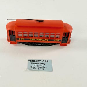 May include: A bright orange model trolley car with black trim, featuring the text 137 BROADWAY 137 on its side and a black roof pole. A small rectangular label is present with the text TROLLEY CAR Broadway POWERED Gay Nineties T214G:798.