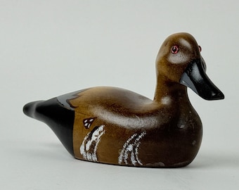 May include: A wooden duck figurine with a brown body, black tail, and a black beak. The duck has red eyes and white painted details on its side. The figurine is a decorative item.