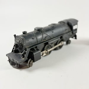 May include: A black toy train with detailed features, including a smokestack, wheels, and a front cowcatcher. The model train is set against a white background, showcasing its classic design. This miniature train is a collectible item.