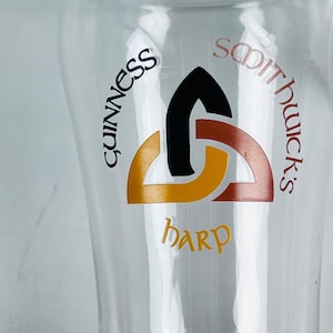 May include: A clear pint glass with the Guinness logo. The logo features a black, orange, and brown design with the words "Guinness", "Smithwick's", and "Harp" printed on the glass.