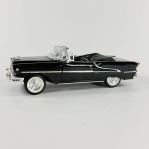 May include: A black convertible car model with white-walled tires and chrome accents. The miniature vehicle has a detailed interior and a clear windshield. The car is a classic design, likely from the mid-20th century.