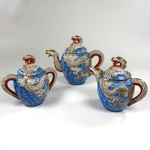 May include: A three-piece porcelain tea set featuring a teapot, a sugar bowl, and a creamer. Each piece is decorated with a vibrant blue background, gold-trimmed dragon motifs, and red accents on the handles, spouts, and lids with dragon finials.