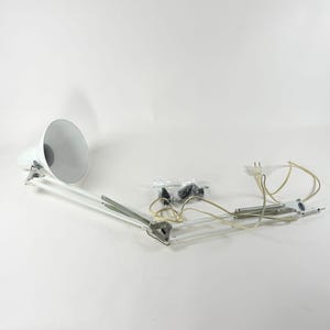 May include: A white architect lamp with an adjustable articulated arm, a wide conical shade, and a long power cord with a two-prong plug. The lamp includes a separate small plastic bag containing black mounting hardware and metal components.