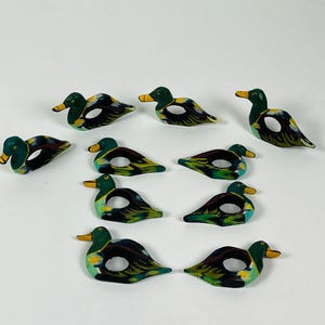 May include: A collection of ten hand-painted duck-shaped napkin rings. Each ring features a green head with a yellow beak, a black and white body, and a circular opening for a napkin. The ducks are arranged on a white surface.