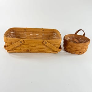 May include: Two woven baskets. One is rectangular with a handle on each side. The other is round with a handle. Both are made of light brown wood and have a clear plastic liner.