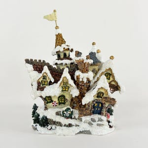 May include: A detailed Christmas village figurine with snow-covered roofs and chimneys. The miniature houses have small windows and doors, with a sign that reads "Frost Bite Clinic". A small flag sits atop one of the towers.