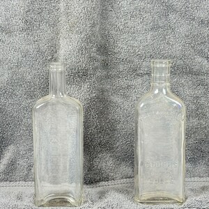 May include: Two clear glass bottles with rectangular bodies and narrow necks. One bottle has the text "FOR COUGHS" and "GOLL'S" embossed on the front. The bottles are empty and sit against a gray, textured background.