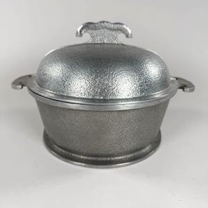 May include: A silver-colored metal casserole dish with a textured surface. The dish has a domed lid with a decorative handle and side handles. The dish is likely used for cooking and serving food.