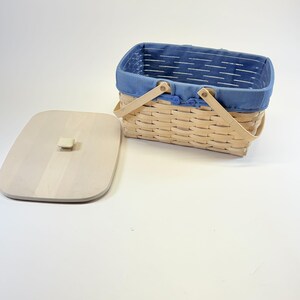 May include: A rectangular picnic basket with a light-coloured woven body and a blue fabric lining. The basket has a wooden handle and a matching wooden lid. The lid is off to the side, revealing the basket's interior.
