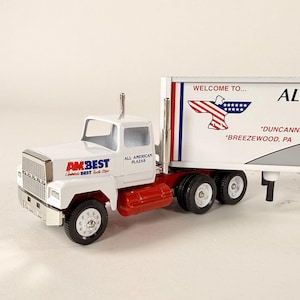 May include: A white AMBEST semi-truck with a red fuel tank and wheels. The trailer has the text "WELCOME TO... ALL AMERICAN PLAZAS" and an eagle graphic. The truck is a model.